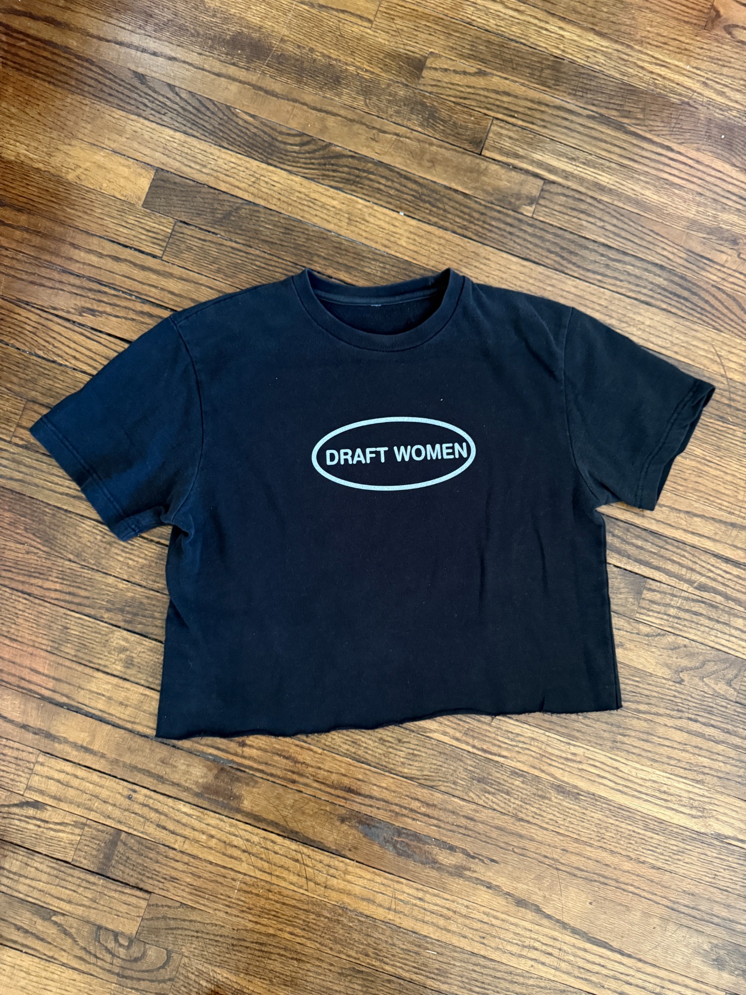 Black Draft Women oval tee on wood floor.